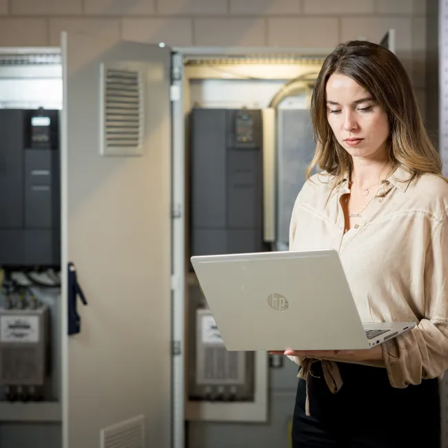 Vrouwelijke engineer met laptop in een technische ruimte na het volgen van de opleiding Hogere installatietechniek voor werktuigkundige installaties fundamentals (HIT-W fundamentals) bij Habeo+.